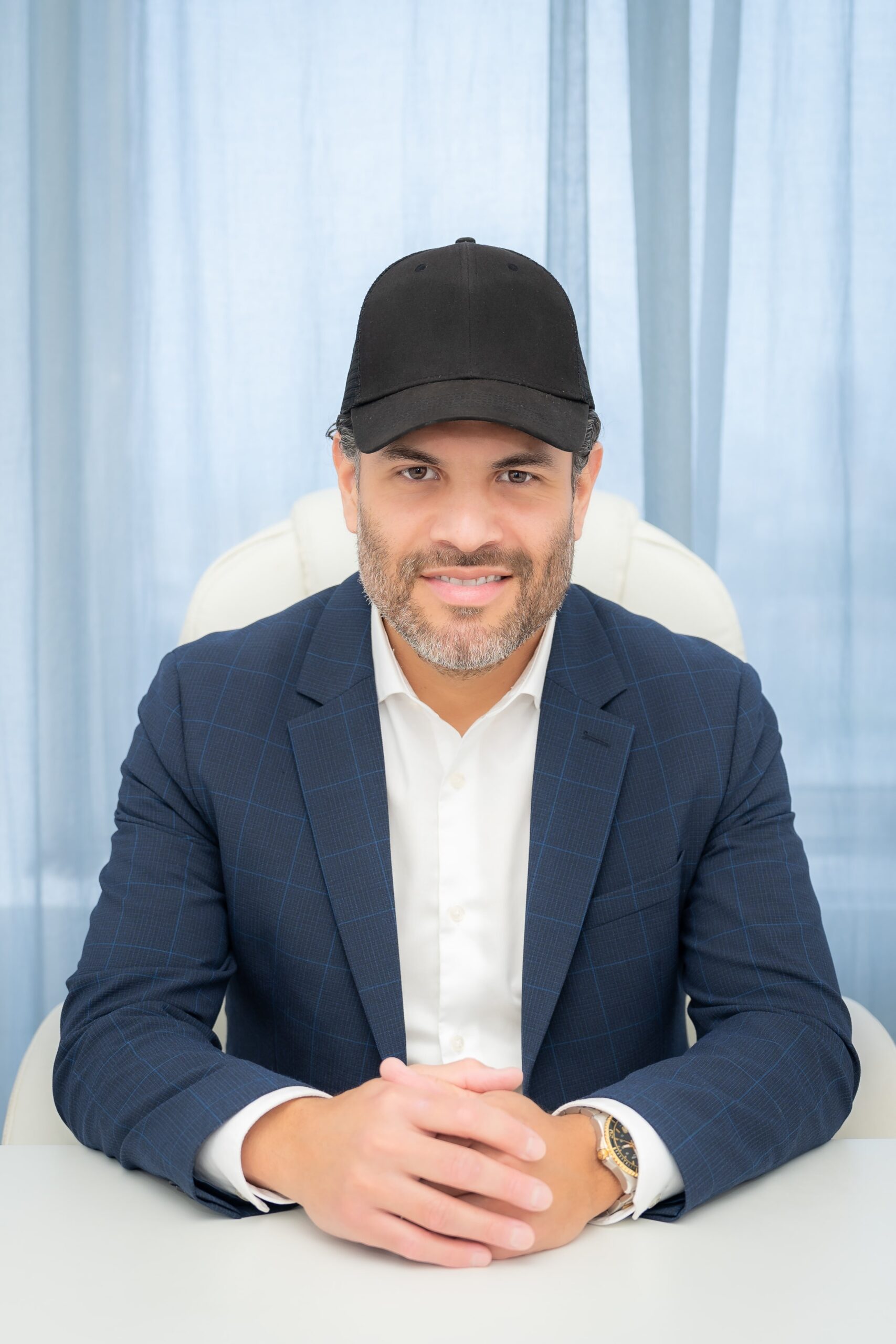 A man dressed in a suit sits at a desk with his hands gently clasped, radiating a sense of calm and balance. His black cap adds a touch of style, while the soothing blue curtains behind him evoke thoughts of tranquility and inner wellness, reminiscent of the serene practices that support gut health.