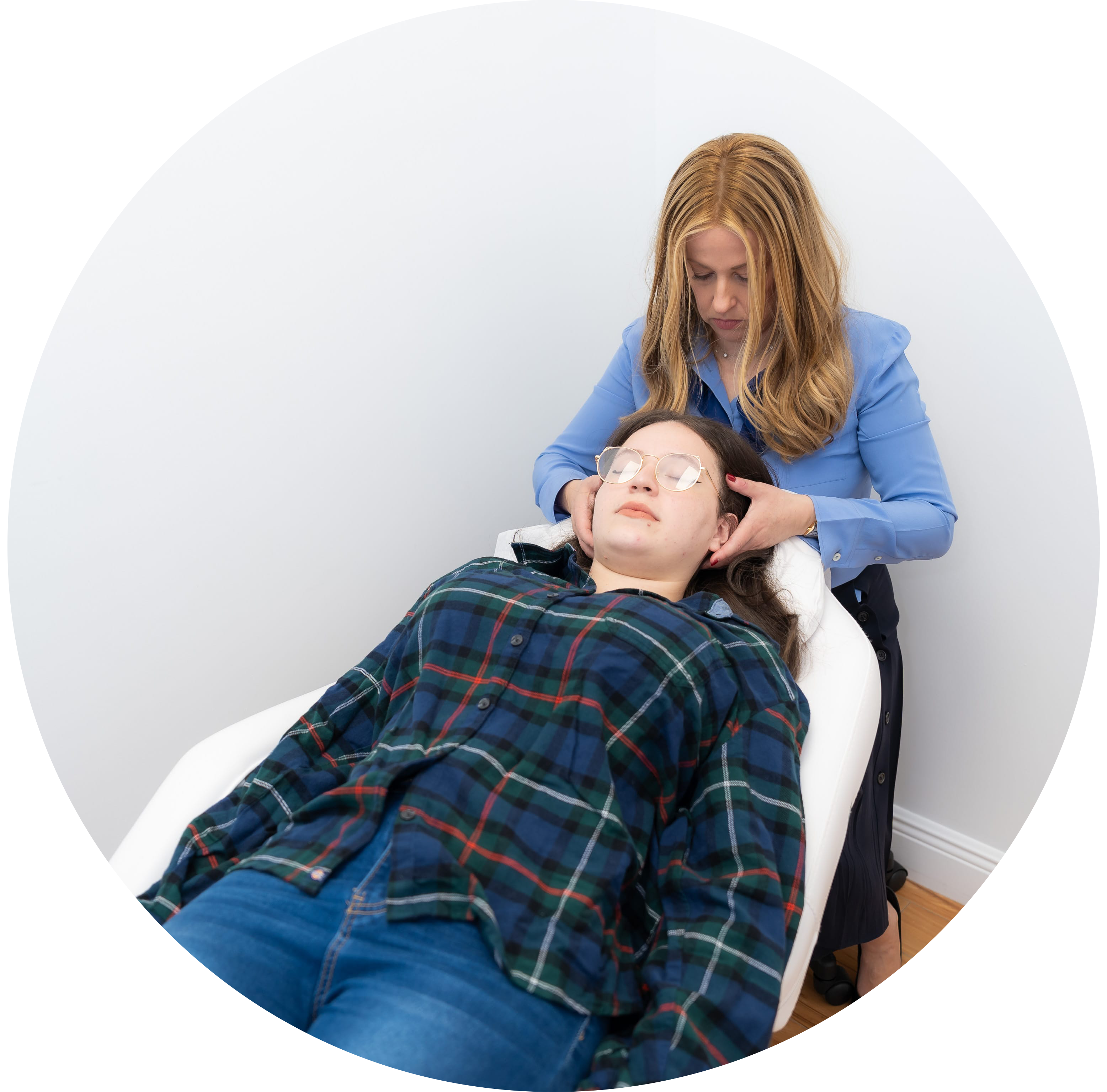 In a tranquil, white-walled room, a woman in a blue shirt carefully supports another woman's head as she relaxes in a comfortable reclining chair, promoting an atmosphere of calm and well-being for better digestion and gut health.