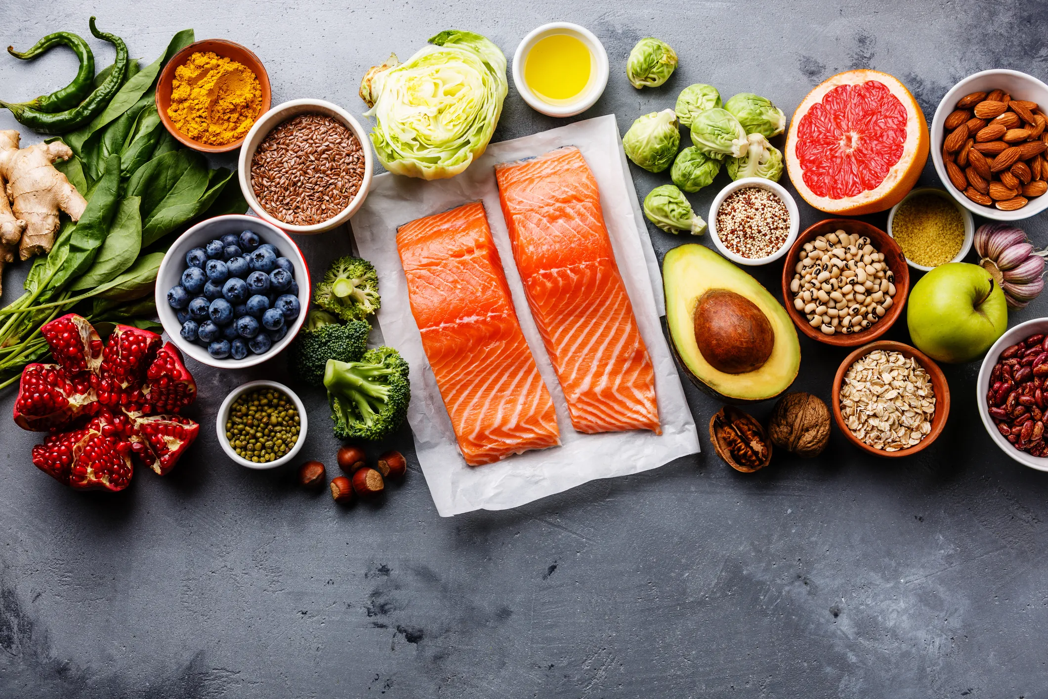 A variety of gut-friendly foods, including fiber-rich fruits and vegetables, omega-3 packed salmon fillets, prebiotic nuts and seeds, and anti-inflammatory spices, arranged on a gray surface.