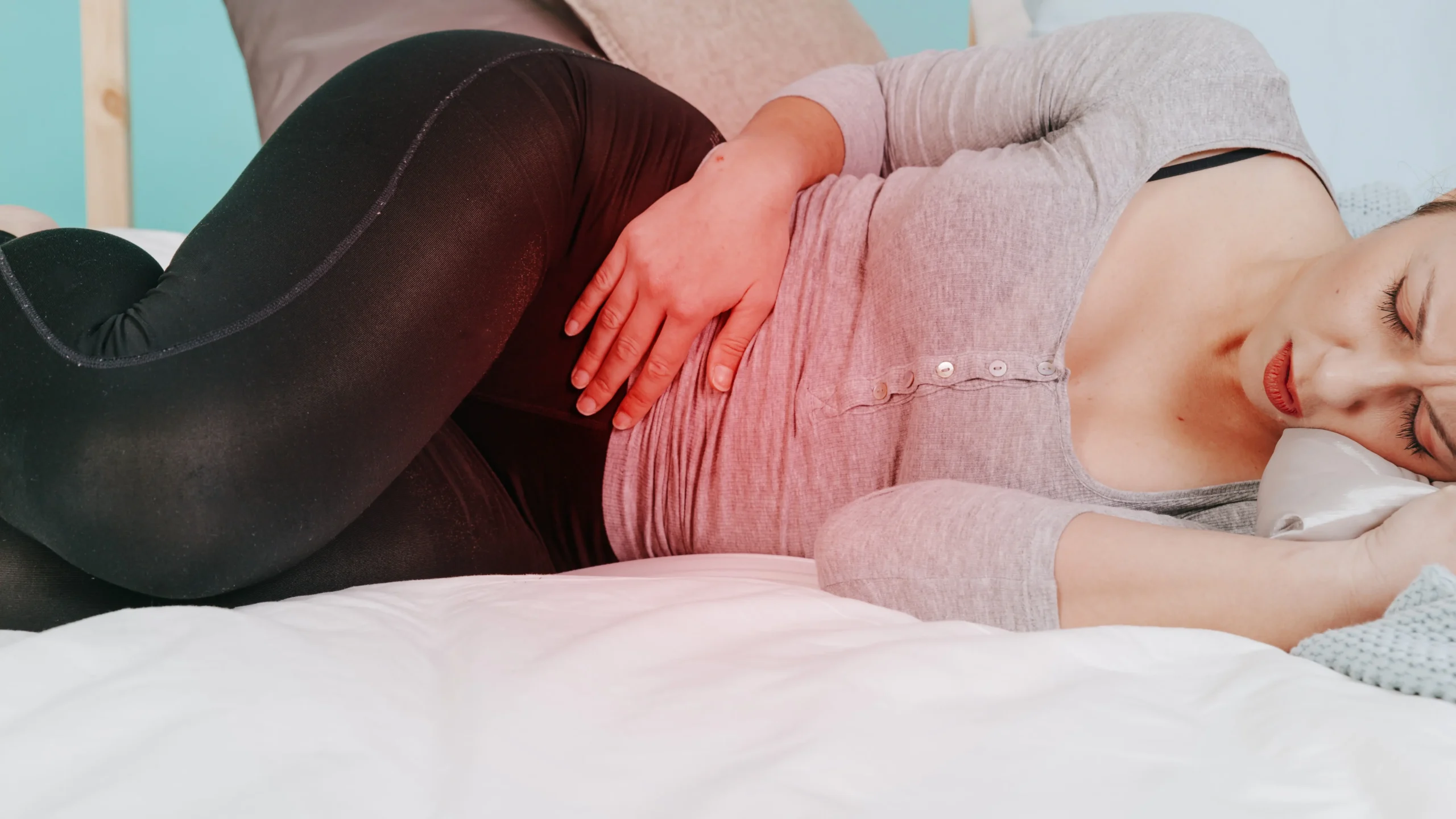 A person lying on their side in bed, indicating signs of digestive discomfort with a hand gently resting on their abdomen. They are dressed in a comfortable gray top and black leggings, suggesting an awareness of the importance of relaxation and comfort for gut health.