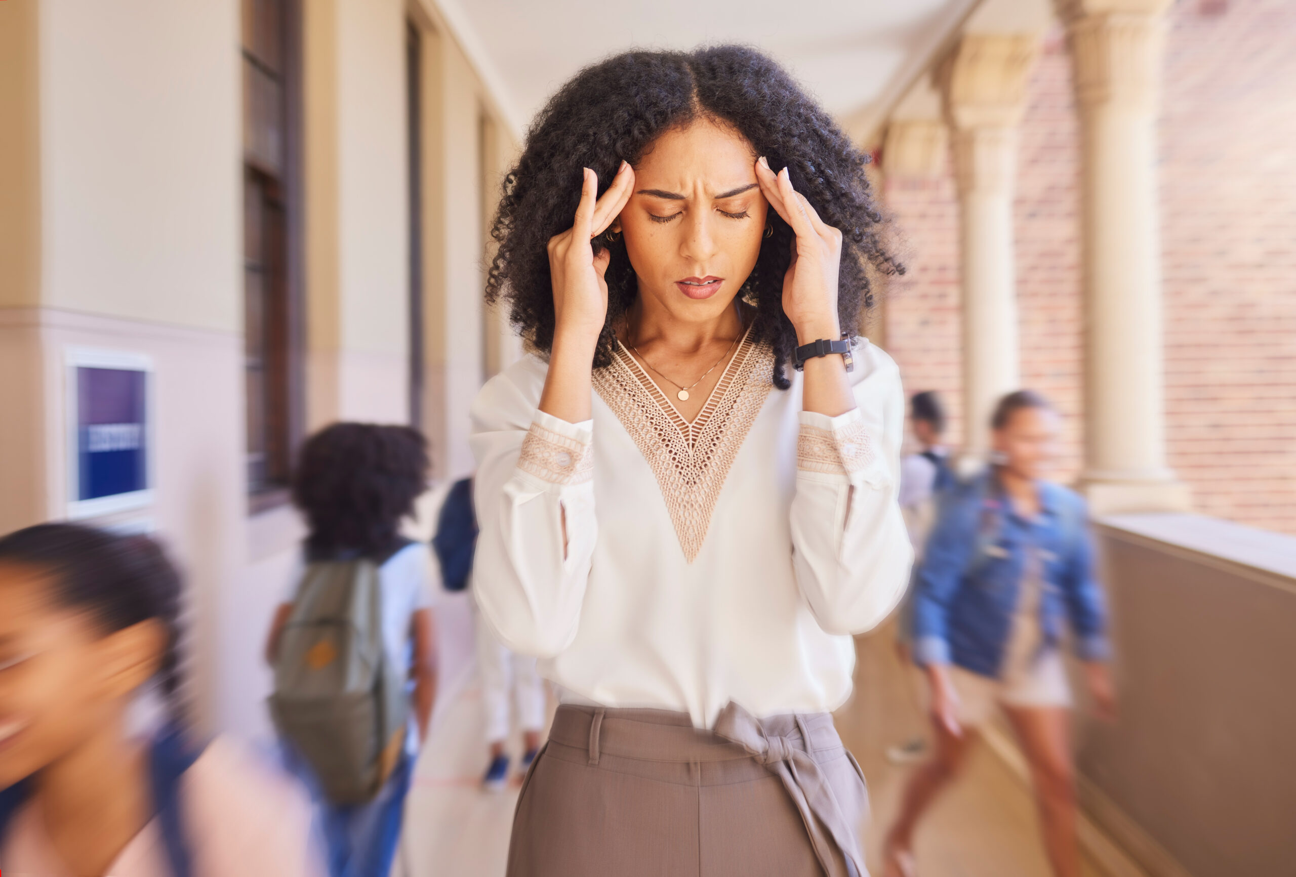 A woman stands in a hallway, gently massaging her temples with her fingers and looking distressed, as people bustle around her.