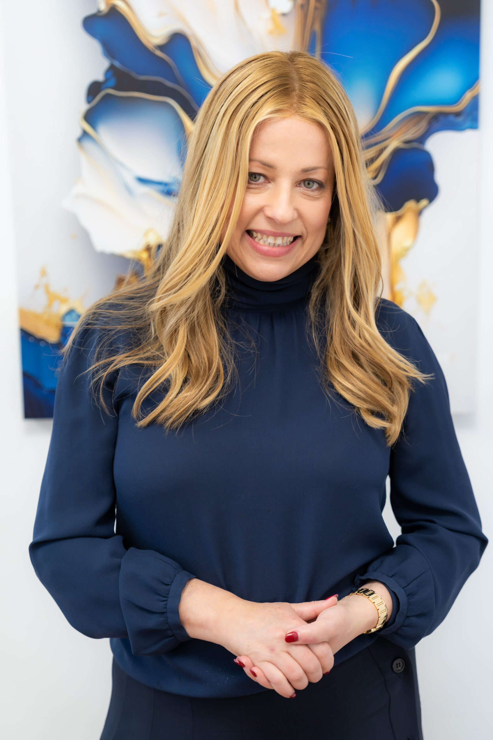 A woman with long blonde hair and a vibrant smile, standing confidently in front of abstract blue and gold art, exuding wellness.