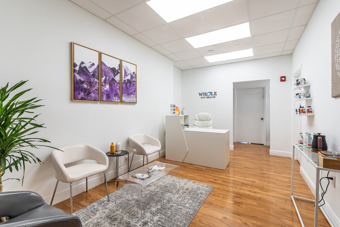 A clean, bright reception with a white desk, chairs, plant, gut health products on shelves, and purple mountain wall art.
